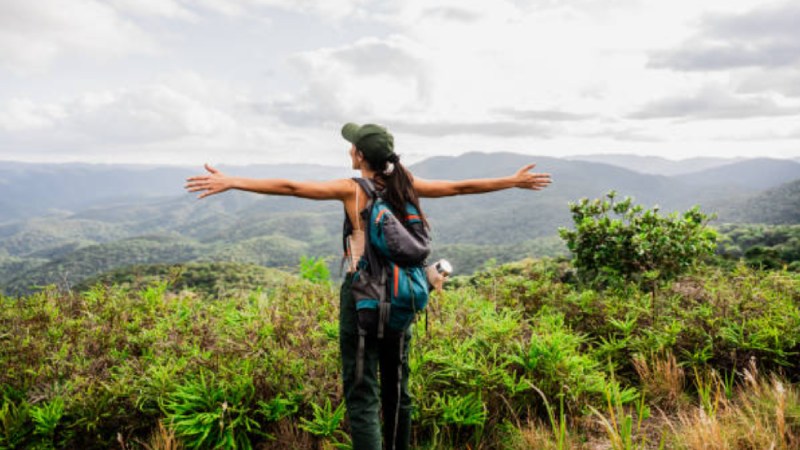 Pessoa com mochila de trilha abre os braços diante de uma vista panorâmica da Mata Atlântica